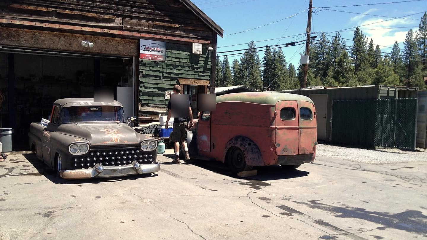 The Ward & Sons shop, a weathered wood and green-shingle building with the bay door open, vintage trucks out front and pines behind